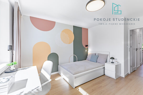 Modern student dorm room with a pastel geometric accent wall, white bed, desk, and nightstand, bathed in natural light west-facing window.