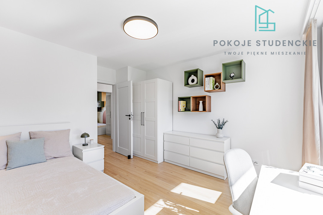 Bright, minimalist student bedroom with white furniture, bed, wardrobe, chest, and wall shelves; logo and text in the corner reading 'POKOJE STUDENCKIE'.