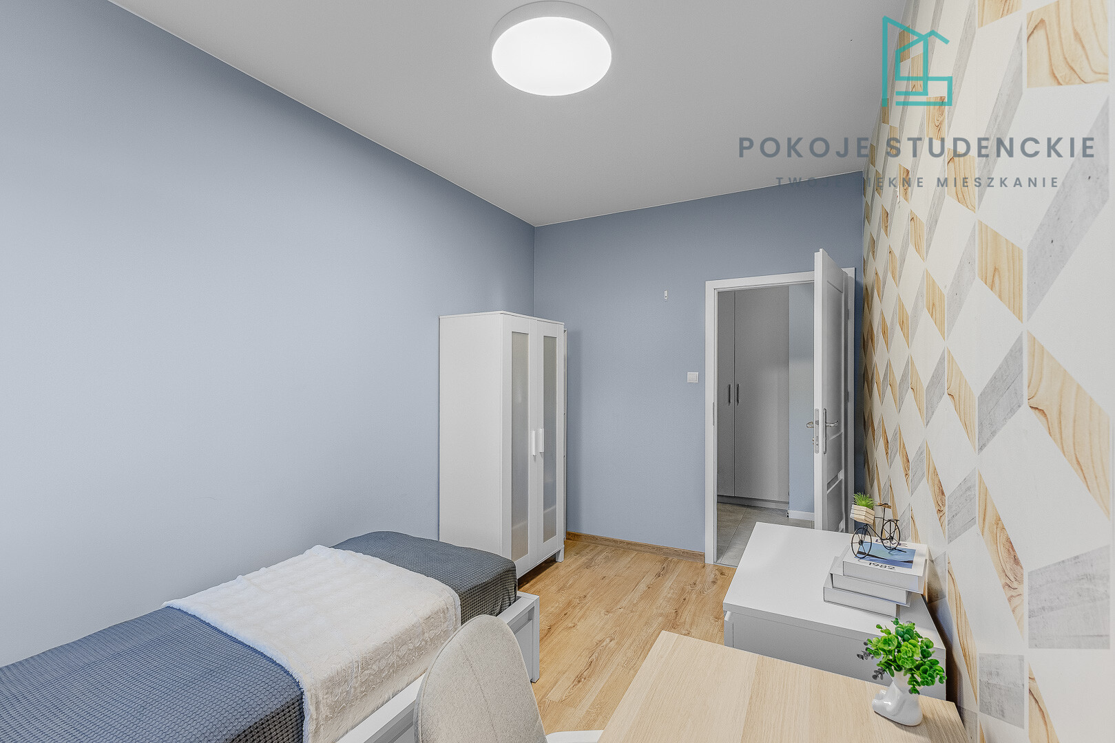 A small modern student room with blue walls, a single bed, white wardrobe, desk, and a geometric wood-pattern accent wall; doorway to another room nearby.
