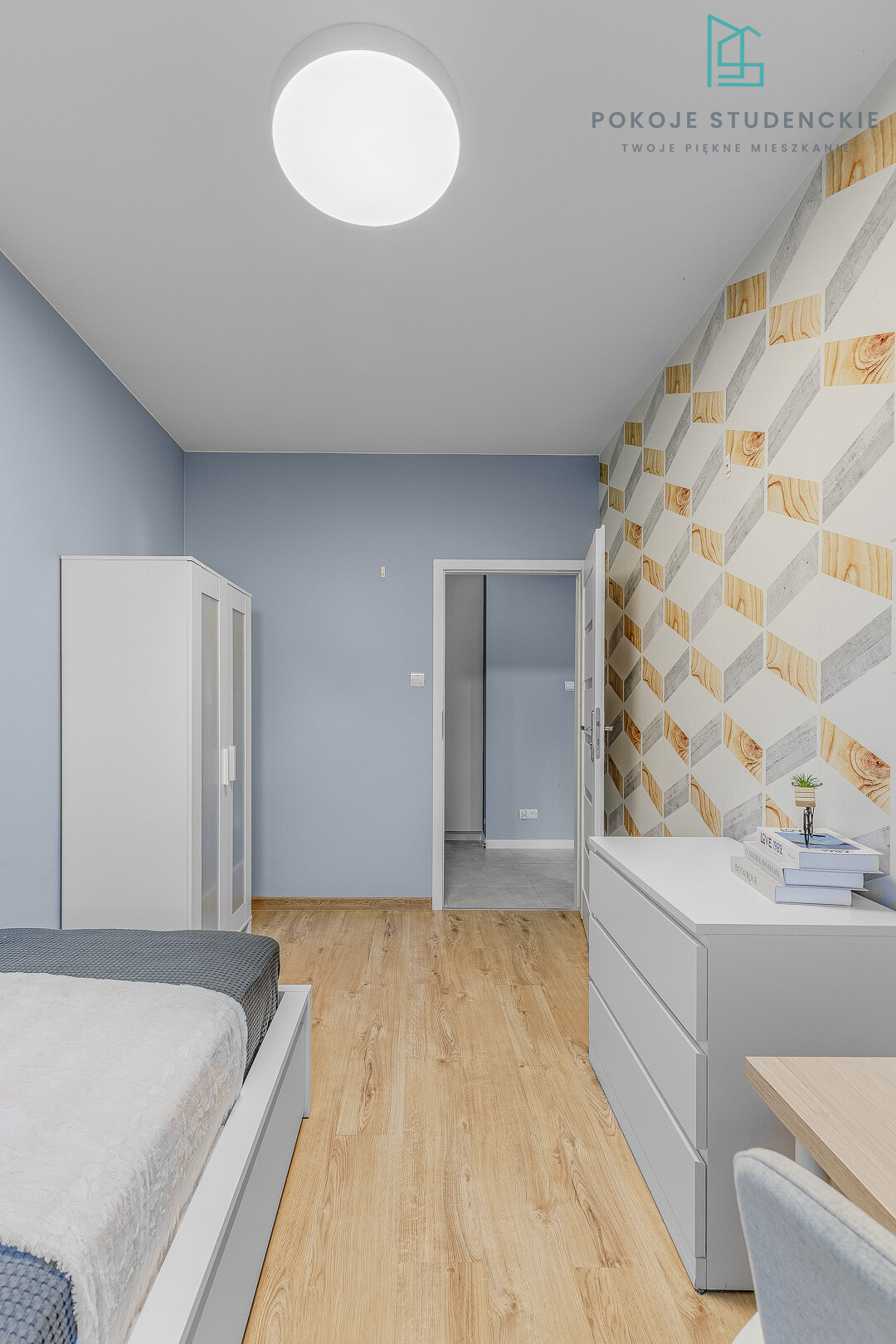 Modern student dorm room with blue walls, white wardrobe and dresser, wood floor, and geometric wallpaper on the right.