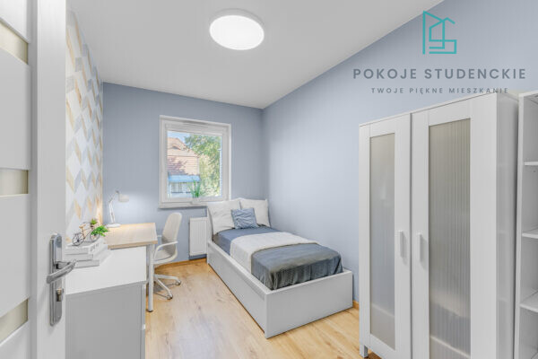 Small student bedroom with light blue walls, a single bed, desk and chair, white wardrobe, and a window showing houses outside.