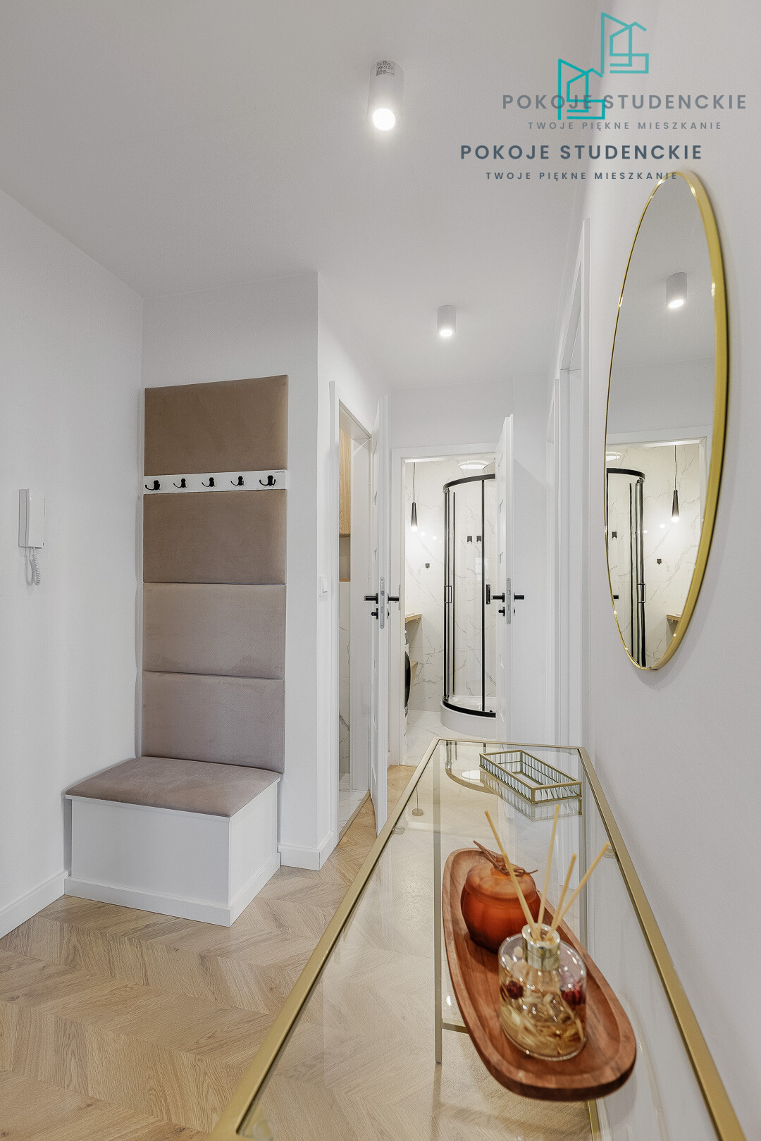 Contemporary hallway with a beige upholstered bench and hooks, a glass console table with a diffuser, and a gold oval mirror on white walls.