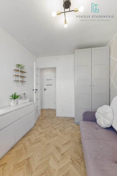 Bright modern dorm room interior with white cabinetry along the left wall, tall gray wardrobe, purple sofa, and a herringbone wood floor.