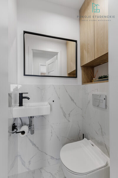 Modern compact bathroom with marble-tiled walls, a wall-mounted white sink, black faucet, and a large black-framed mirror above it, plus wooden cabinetry on the right.