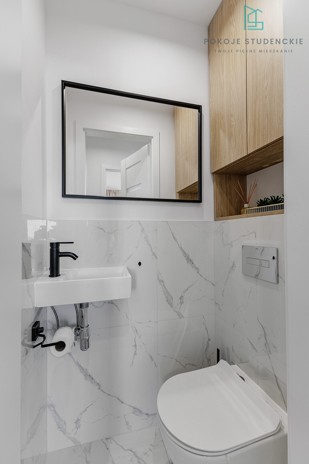Modern compact bathroom with marble-tiled walls, a wall-mounted white sink, black faucet, and a large black-framed mirror above it, plus wooden cabinetry on the right.