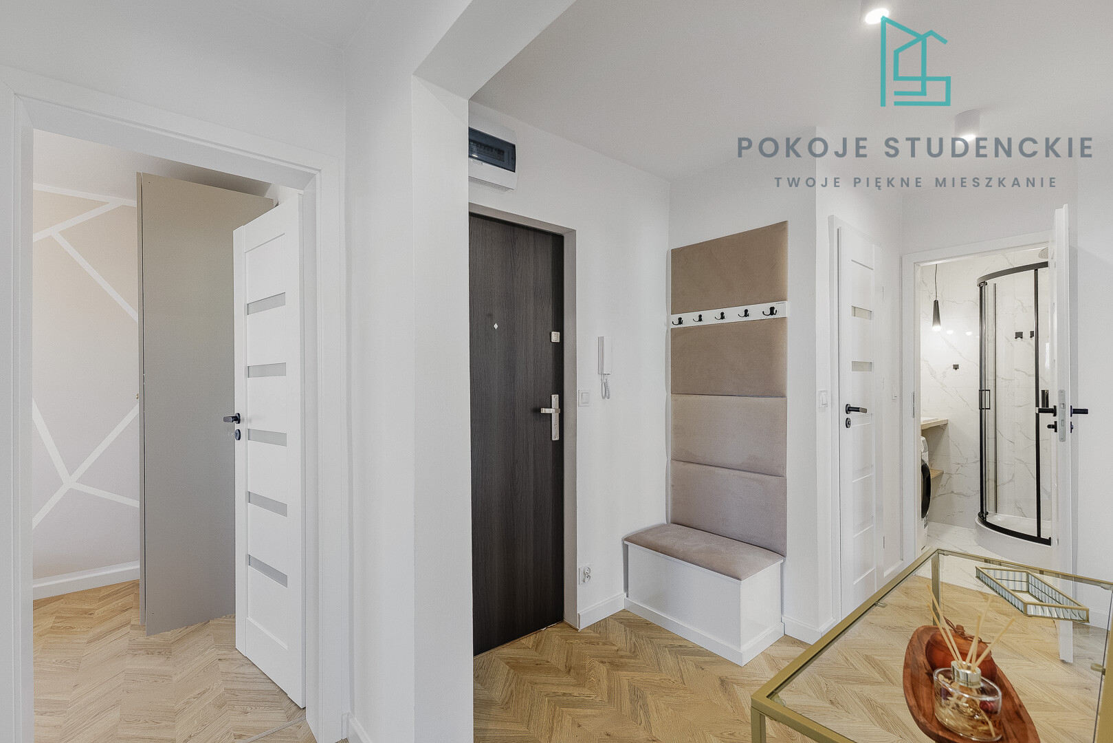 Bright dormitory hallway with white walls, a dark entry door, a padded bench with hooks, and light wood flooring.