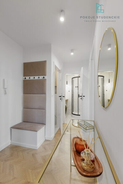 Bright entry hall with a beige padded bench and hooks, a glass console table, and a gold oval mirror; doorway to a marble-tiled bathroom.