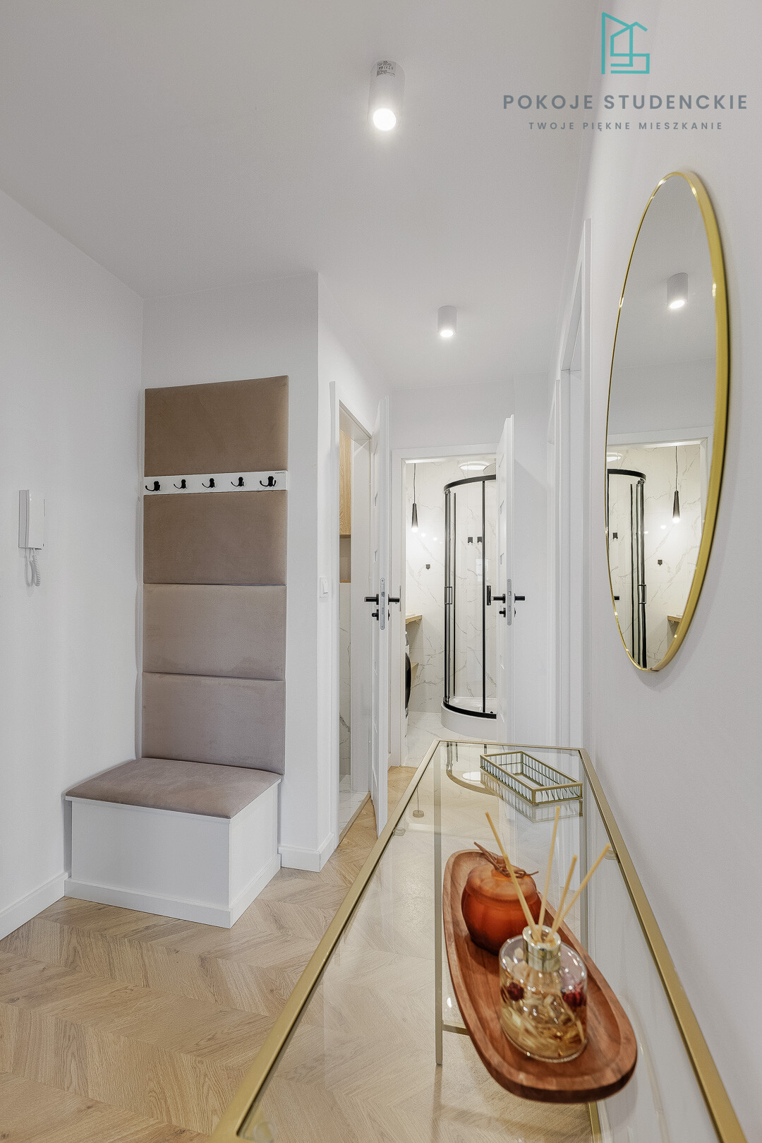 Bright entry hall with a beige padded bench and hooks, a glass console table, and a gold oval mirror; doorway to a marble-tiled bathroom.