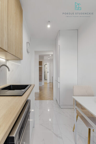 Modern white kitchen with wooden upper cabinets, black cooktop, stainless oven, and marble backsplash; dining table with a beige chair visible to the right.