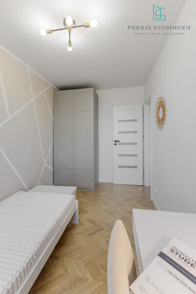 Small modern bedroom with gray wardrobe, white bed, and a white desk; geometric wall design and herringbone wood floor; ceiling light fixture and a logo in the corner.