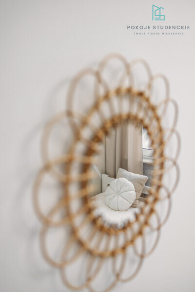 Circular woven mirror on a light wall reflecting a cozy white bed with a round tufted cushion and beige pillow, plus a window with sheer curtains (branding in top-right).