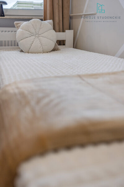 Round, cream tufted pillow on a white textured bedspread in a bright, minimalist bedroom by a window with beige curtains.