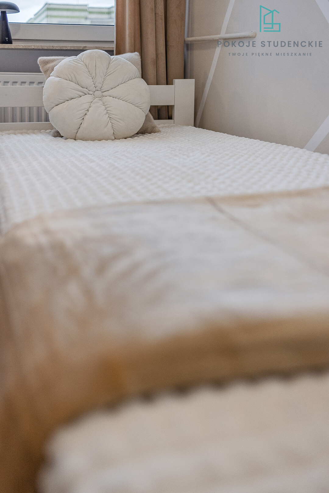 Round, cream tufted pillow on a white textured bedspread in a bright, minimalist bedroom by a window with beige curtains.