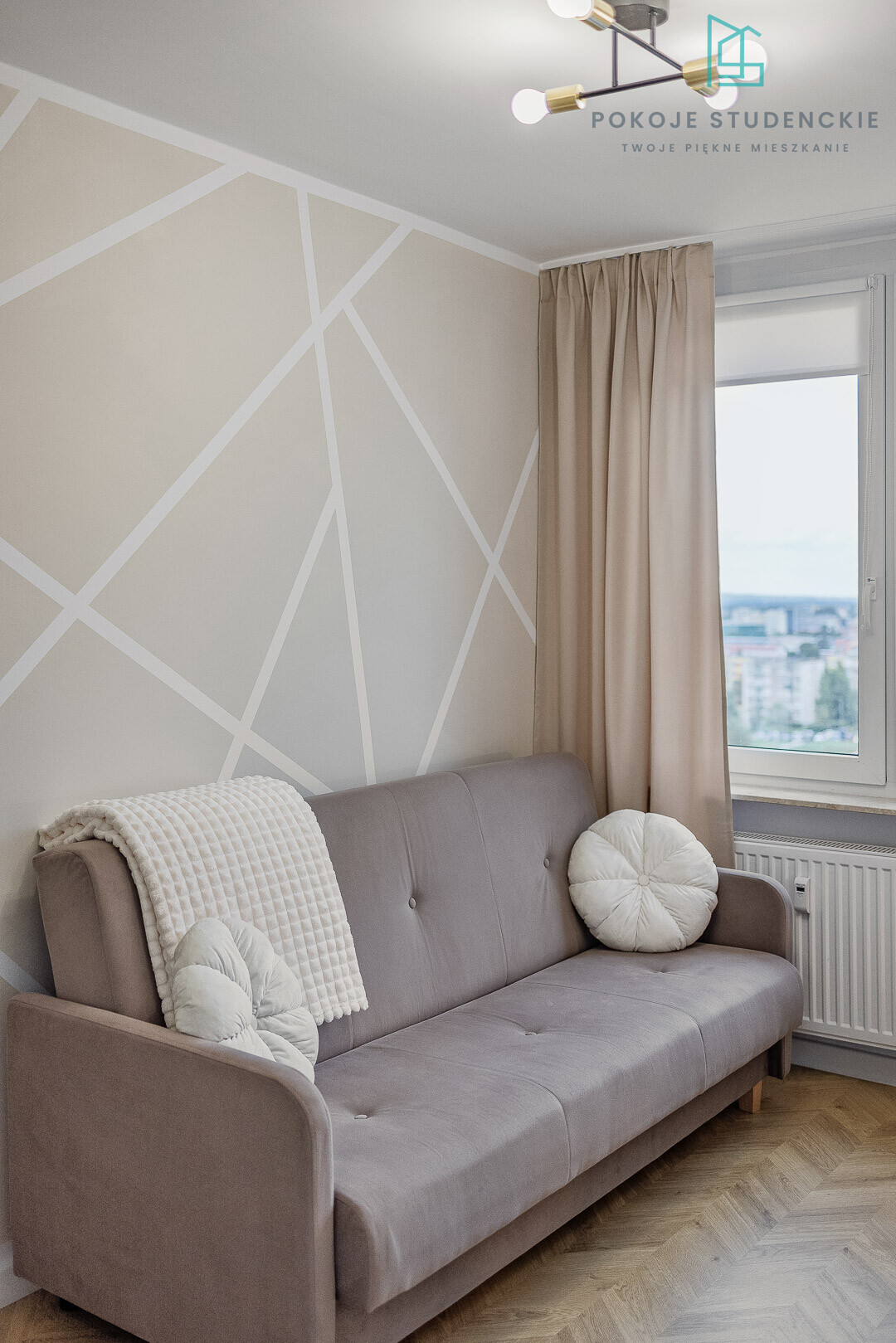 Living room with a gray sofa, cream throw, and round pillows in front of a geometric beige wall near a window with beige curtains.