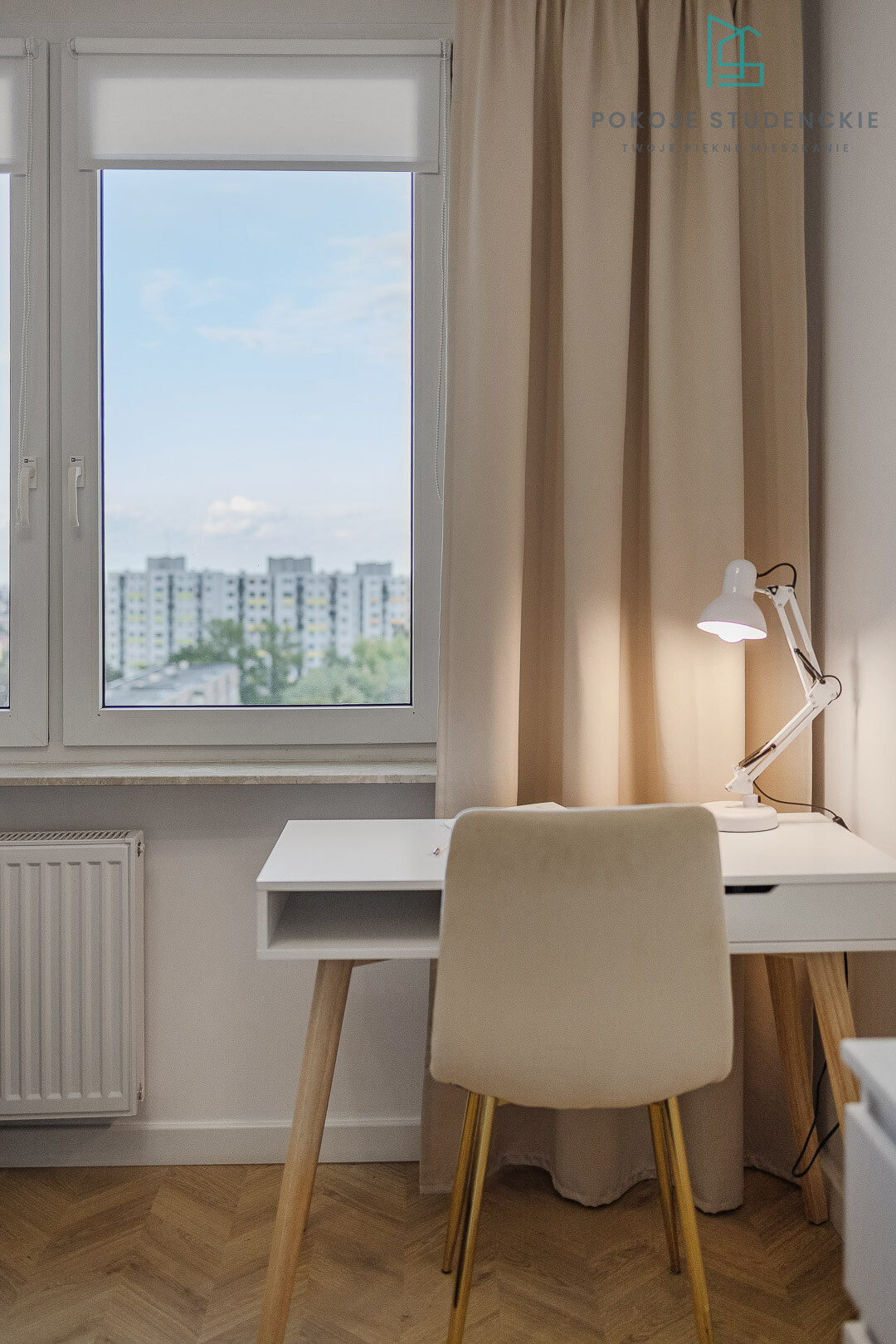 Light-filled study nook with a white desk, beige chair, and adjustable lamp by a large window overlooking a cityscape with beige curtains to the right.