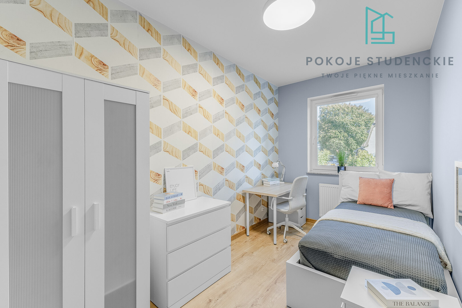 Bright modern bedroom with a geometric patterned accent wall, white wardrobe and dresser, desk with chair, and a single bed by a large window.
