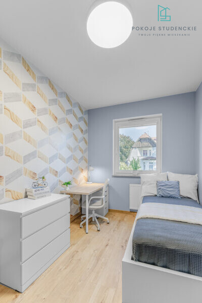 Student bedroom with blue walls, geometric wallpaper, a study desk and chair, white dresser, and a single bed by a window.